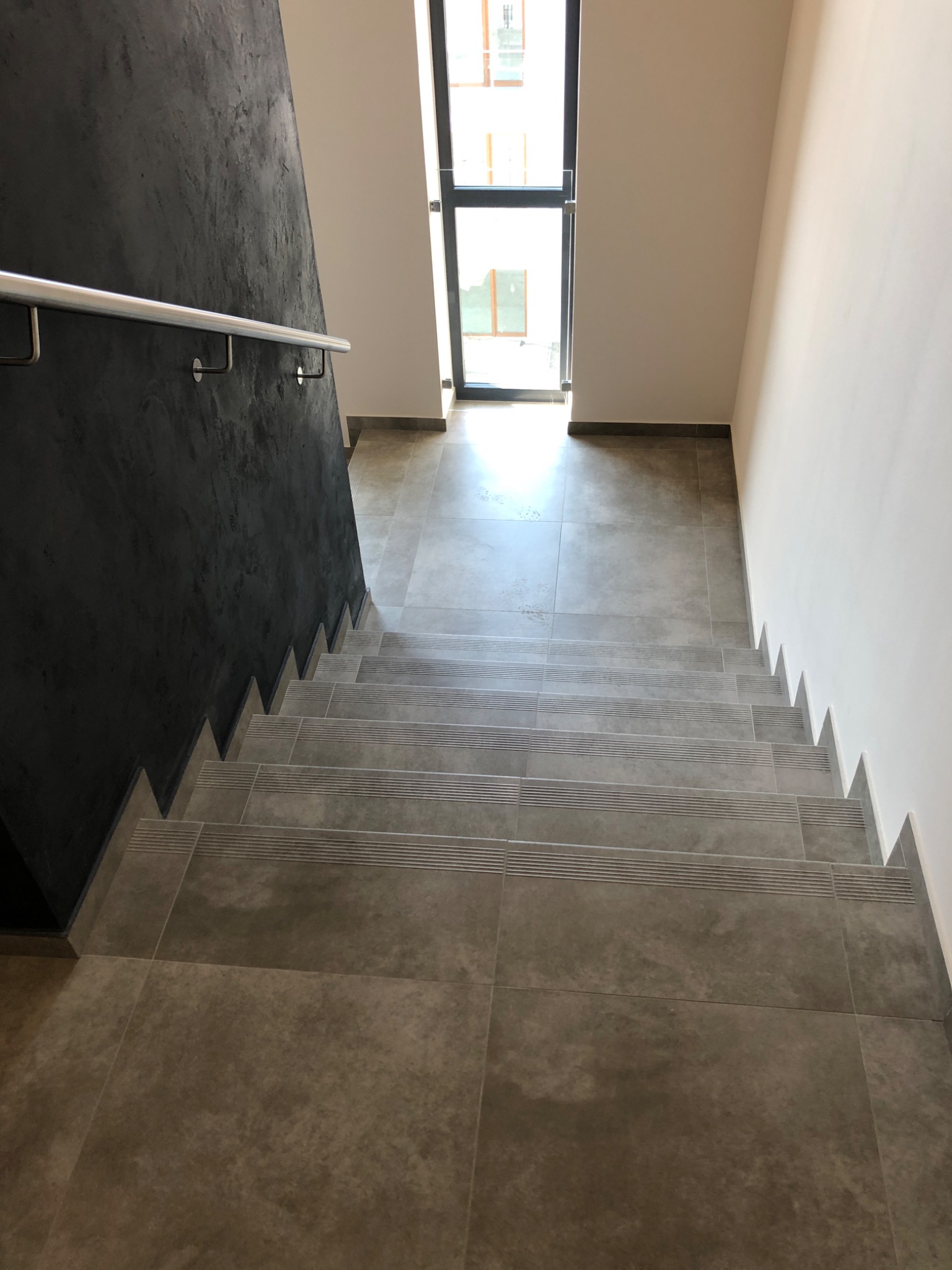 Schody wykończone szarymi płytkami z antypoślizgowymi paskami, czarna ściana z efektem betonu i metalowa poręcz, widok z góry w kierunku okna.