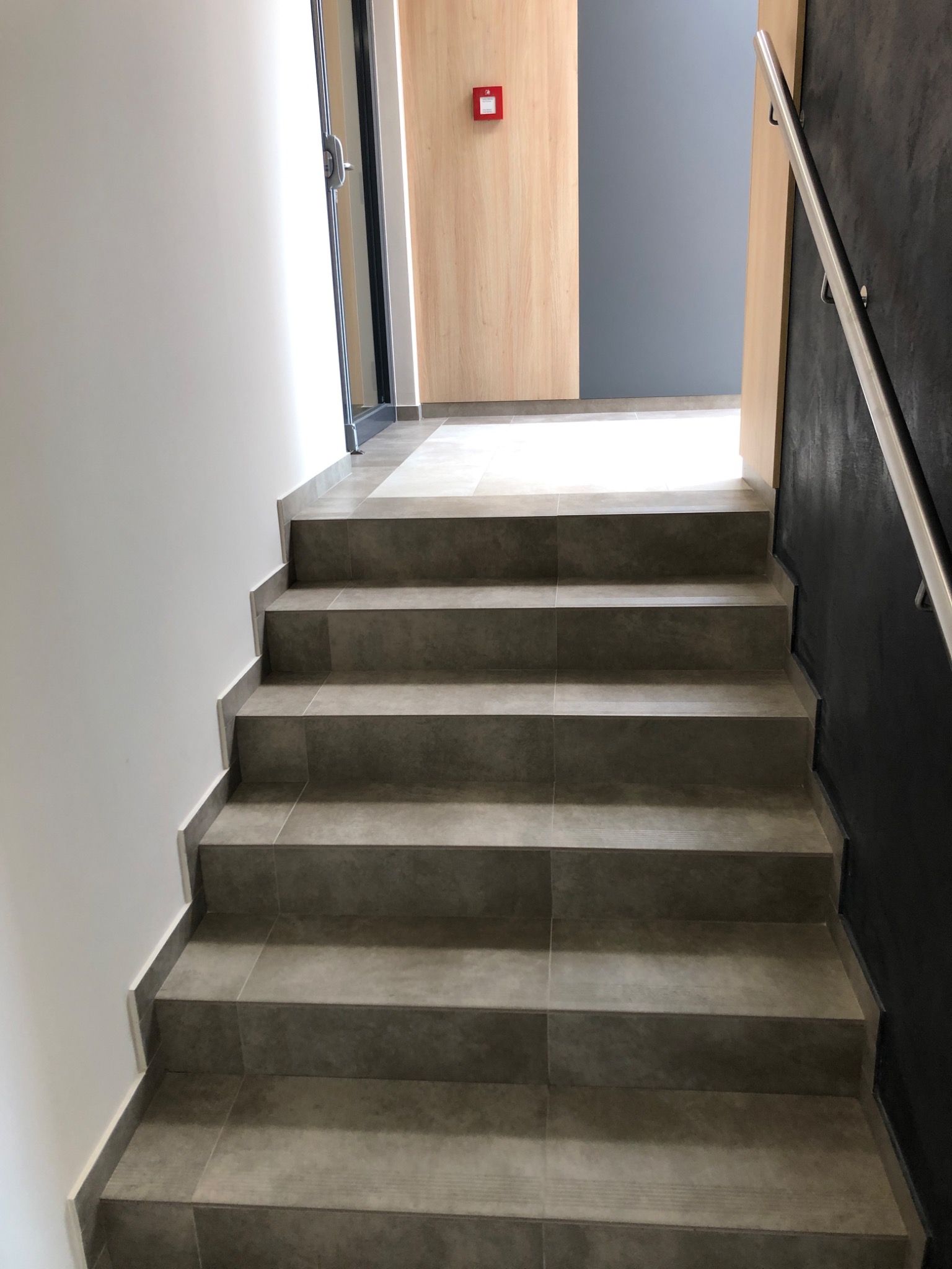 Schody wyłożone szarymi płytkami gresowymi, widok z dołu, z metalową poręczą po prawej stronie i fragmentem drzwi z drewnopodobną okleiną w tle.