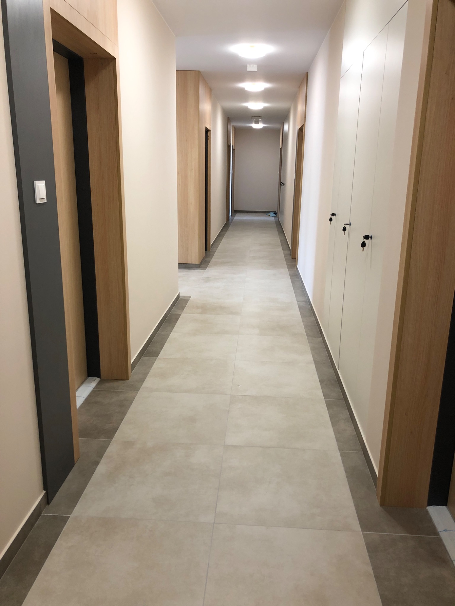Długi korytarz w jasnych barwach z drewnianymi drzwiami i szafami w zabudowie, wykończony płytkami podłogowymi w odcieniach beżu i szarości.