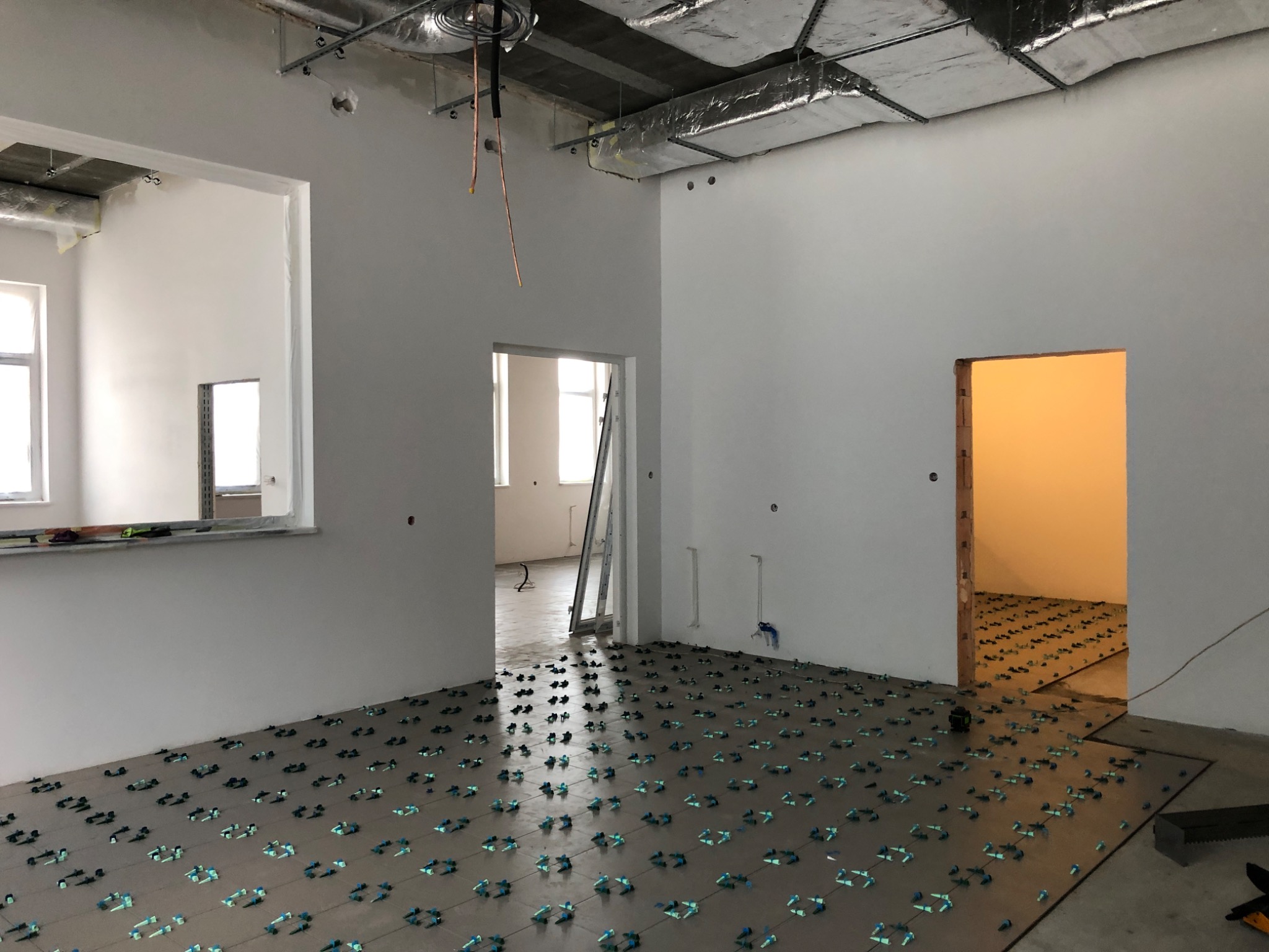 Przygotowanie podłoża pod płytki w dużej przestrzeni biurowej, widoczne poziomice i krzyżyki dystansowe, surowy stan ścian i sufitu z widoczną instalacją wentylacyjną.