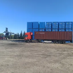 Czerwona ciężarówka z brązowym kontenerem Honeytak na tle ściany z niebieskich kontenerów, w tle wózek widłowy.