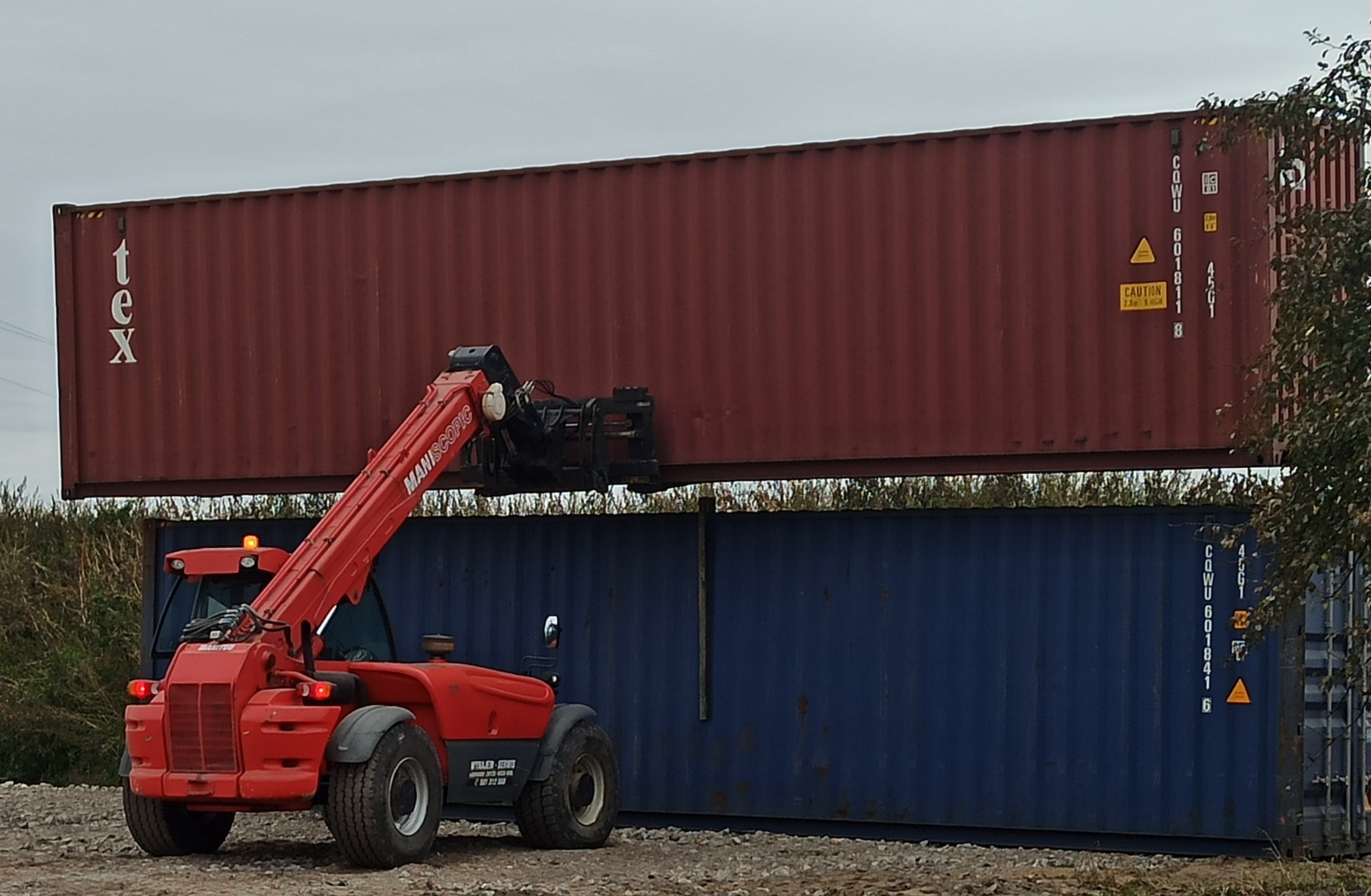 Czerwony wózek widłowy Maniscopic podnosi kontener TEX na tle niebieskiego kontenera.