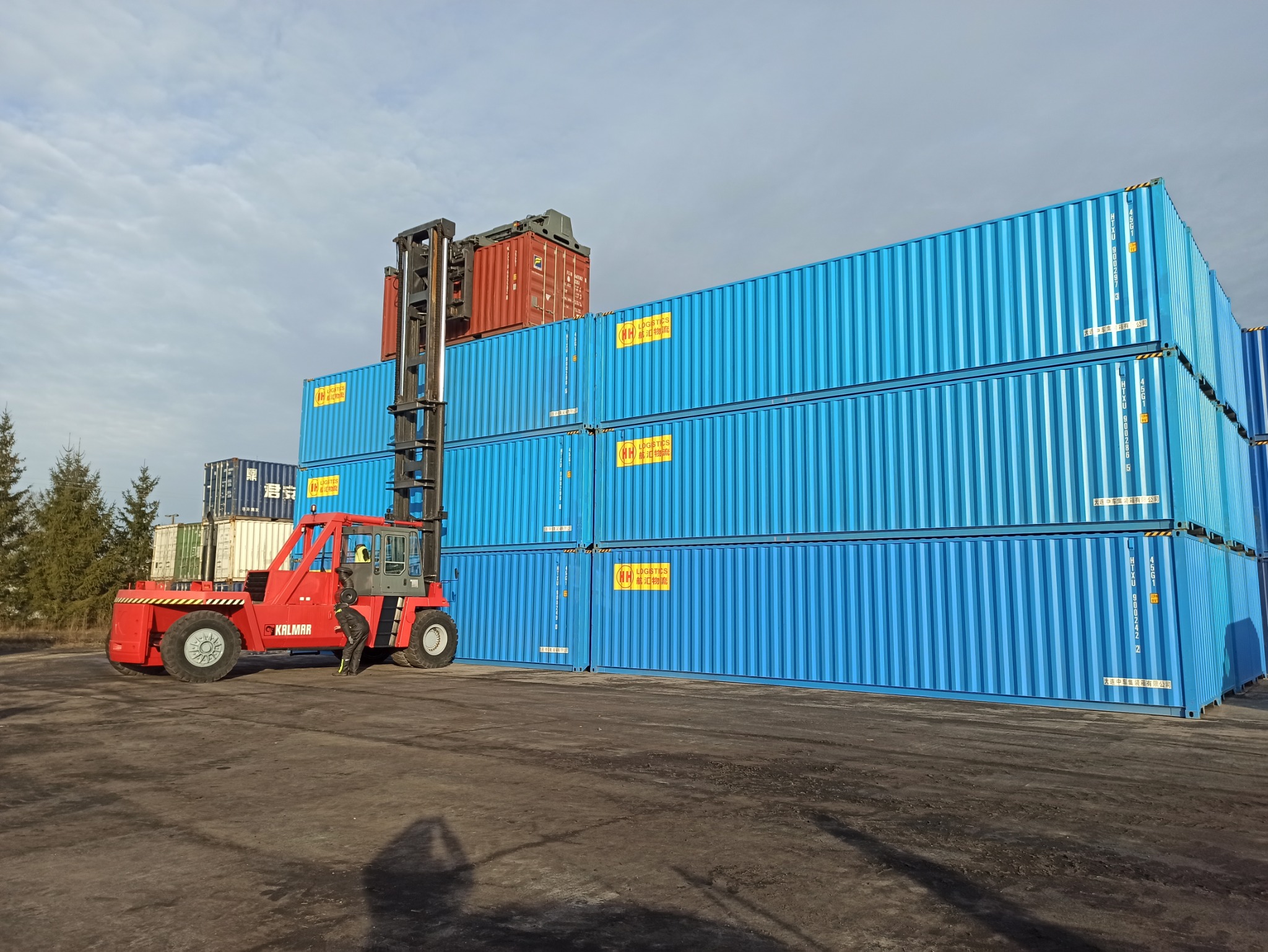 Czerwony wózek widłowy Kalmar podnosi czerwony kontener na stos niebieskich kontenerów z żółtymi naklejkami, widoczne drzewa w tle.