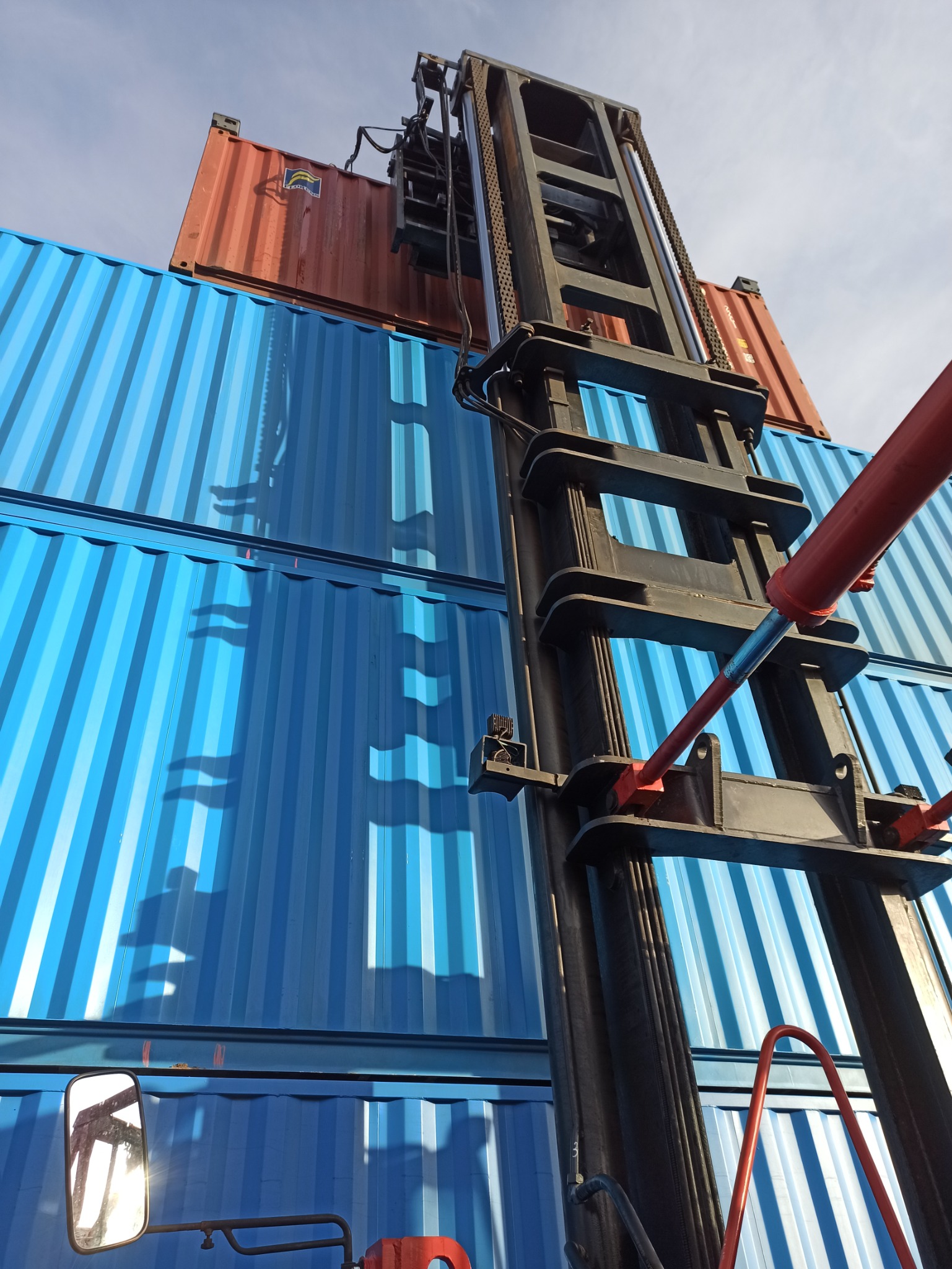 Wózek widłowy podnosi czerwony kontener na stos niebieskich kontenerów, widok z dołu, błękitne niebo w tle.