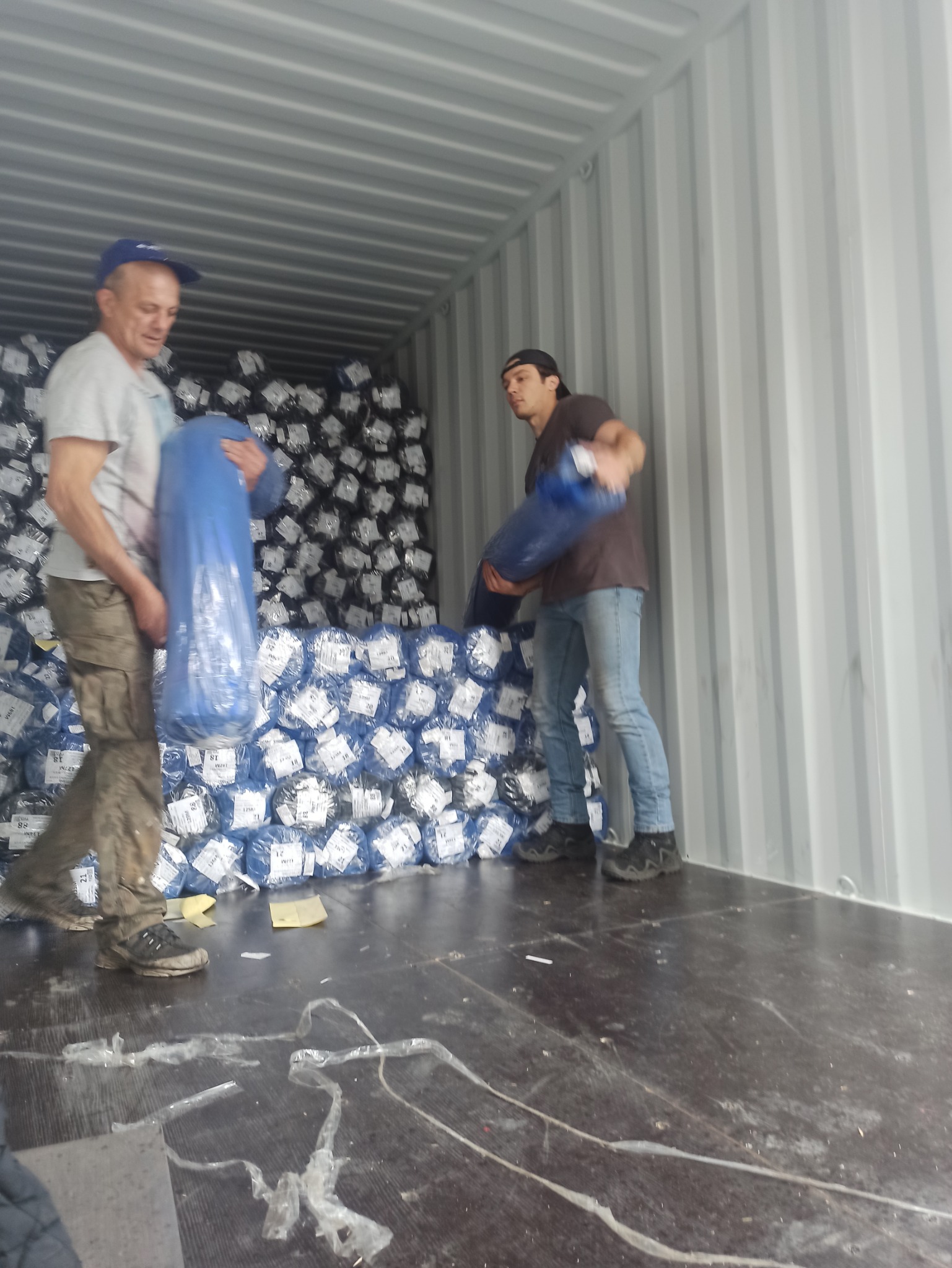 Dwóch mężczyzn rozładowuje kontener z dużą ilością zafoliowanych rolek materiału, widoczne etykiety na rolkach, podłoga kontenera zabrudzona resztkami folii.