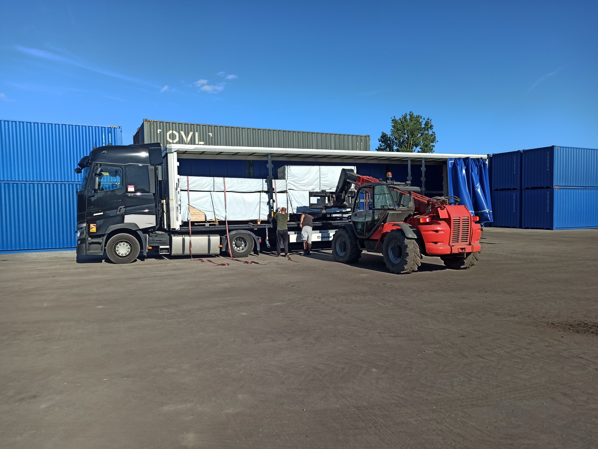 Rozładunek towaru z naczepy ciężarówki za pomocą wózka widłowego teleskopowego na tle niebieskich kontenerów.