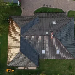 Widok z góry na dom z ciemną, nową dachówką ceramiczną, z widocznymi oknami dachowymi, kominem, trawnikiem i robotem koszącym w rogu.