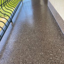 Izolujmy.pl - Błyszcząca, brązowa posadzka balkonu z dekoracyjnym wzorem z drobnych kamyczków, widok na metalową balustradę i zieleń na zewnątrz.