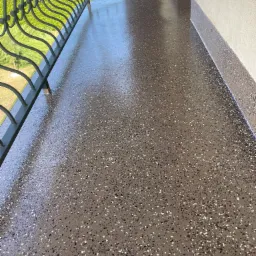 Błyszcząca, brązowa posadzka balkonu z dekoracyjnym wzorem z drobnych kamyczków, widok na metalową balustradę i zieleń na zewnątrz.