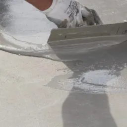 Ręka w roboczej rękawicy rozprowadza szpachlą mokrą, samopoziomującą masę na betonowej powierzchni, tworząc gładką warstwę.