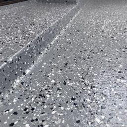 Izolujmy.pl - Szara, wielobarwna, żywiczna posadzka balkonu lub tarasu z widocznym progiem drzwi balkonowych i fragmentem ściany.