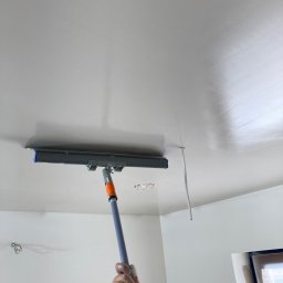 Izolujmy.pl - Aplikacja gładzi na suficie za pomocą szpachli na kiju teleskopowym, widoczna ręka z obrączką i fragment ściany z wystającym kablem.
