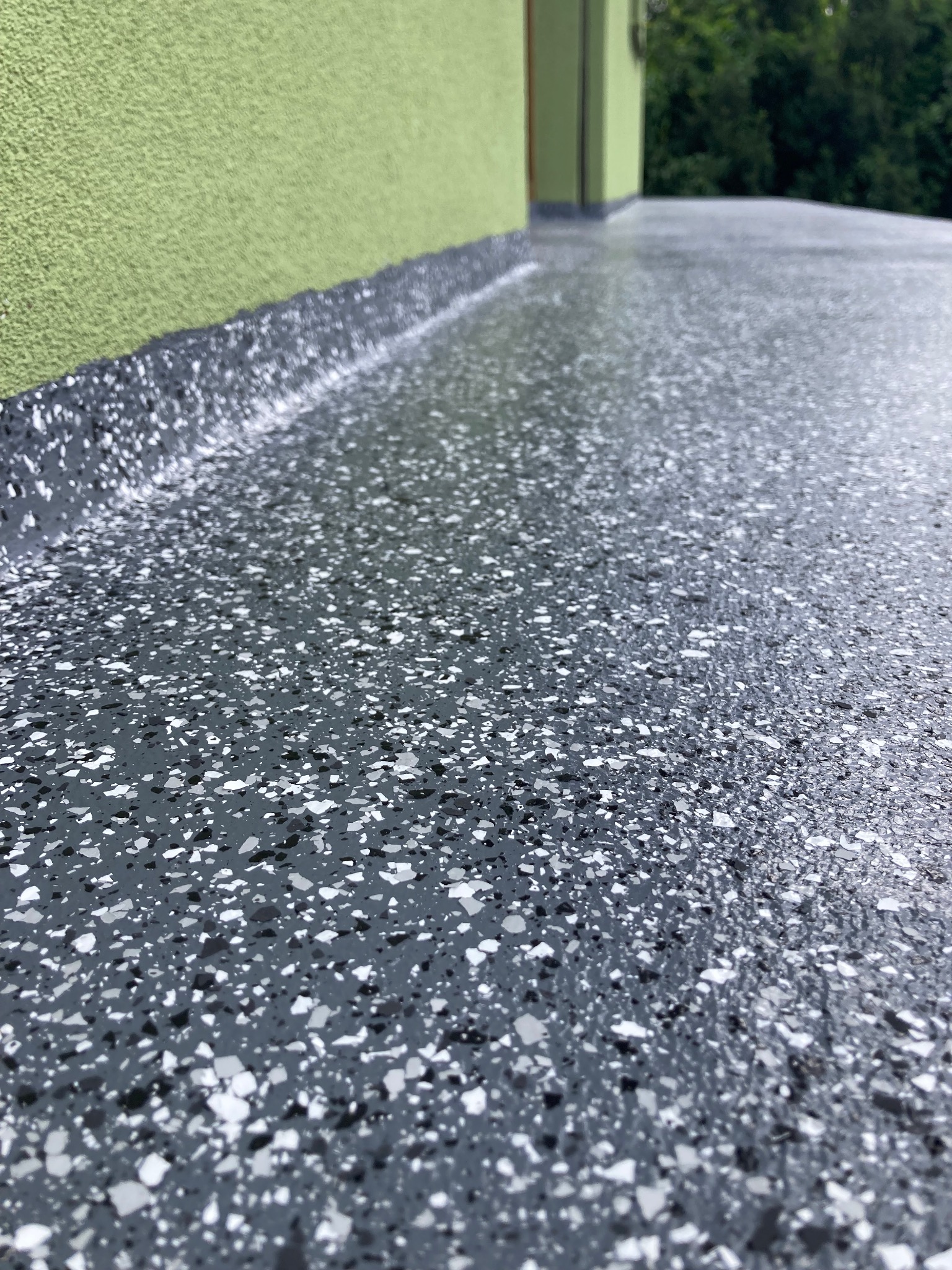 Szara, żywiczna posadzka tarasu lub balkonu z białymi i czarnymi wtrąceniami, widoczna ściana budynku w kolorze zielonym.