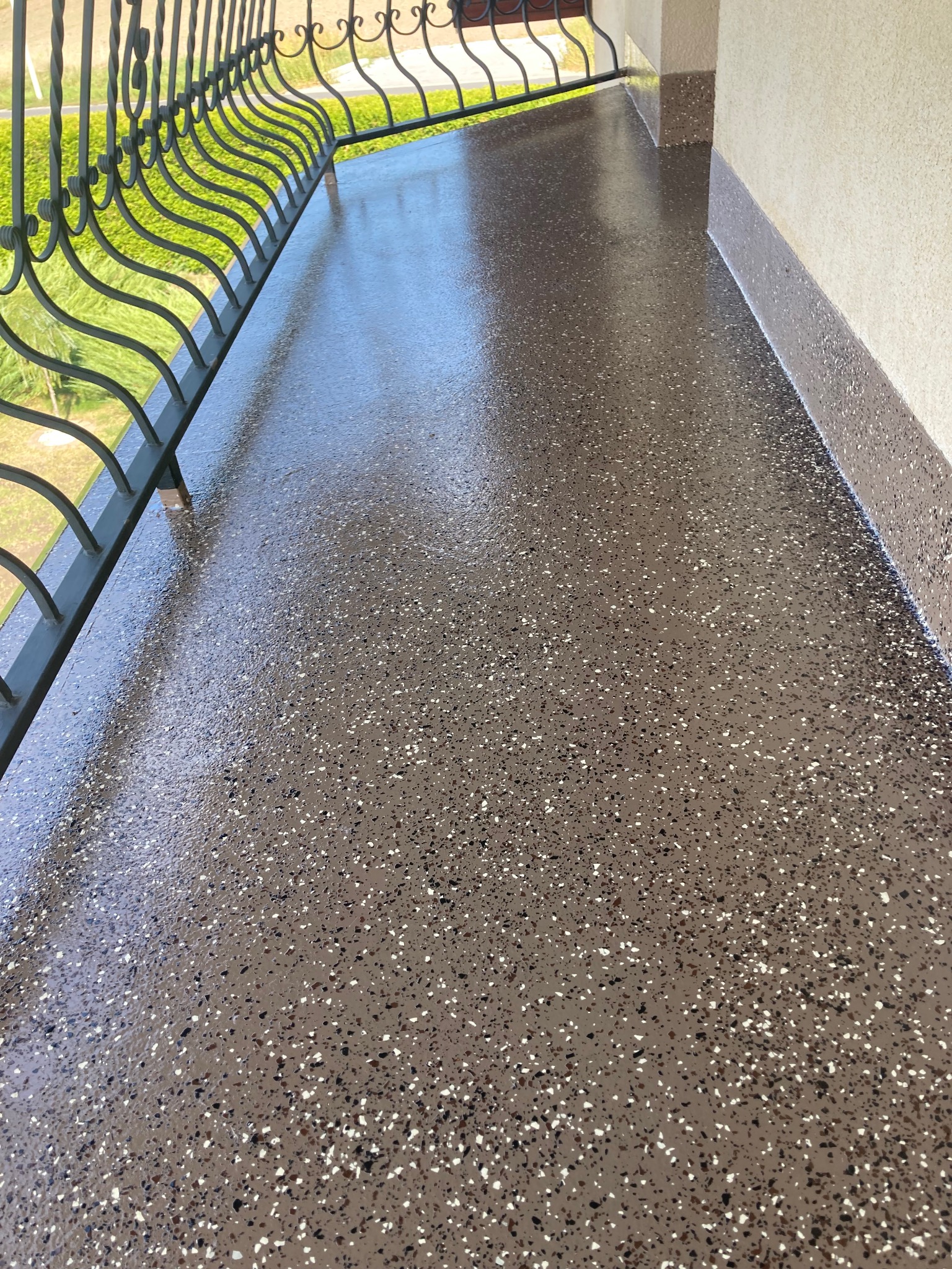 Błyszcząca, brązowa posadzka balkonu z dekoracyjnym wzorem z drobnych kamyczków, widok na metalową balustradę i zieleń na zewnątrz.