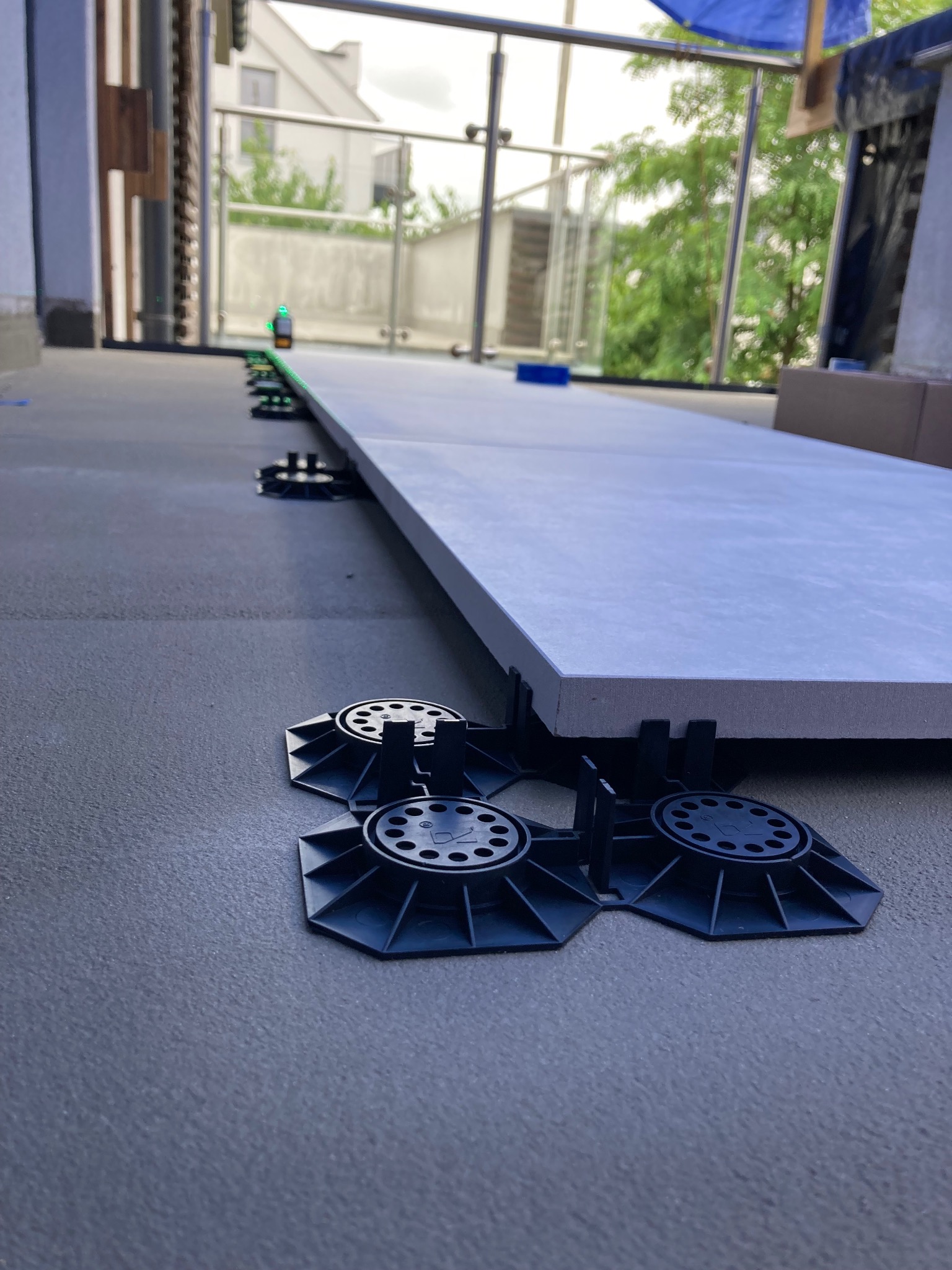 Montaż szarych płyt tarasowych na regulowanych wspornikach z tworzywa, widoczny laser poziomujący, balkon z szklaną balustradą w tle.