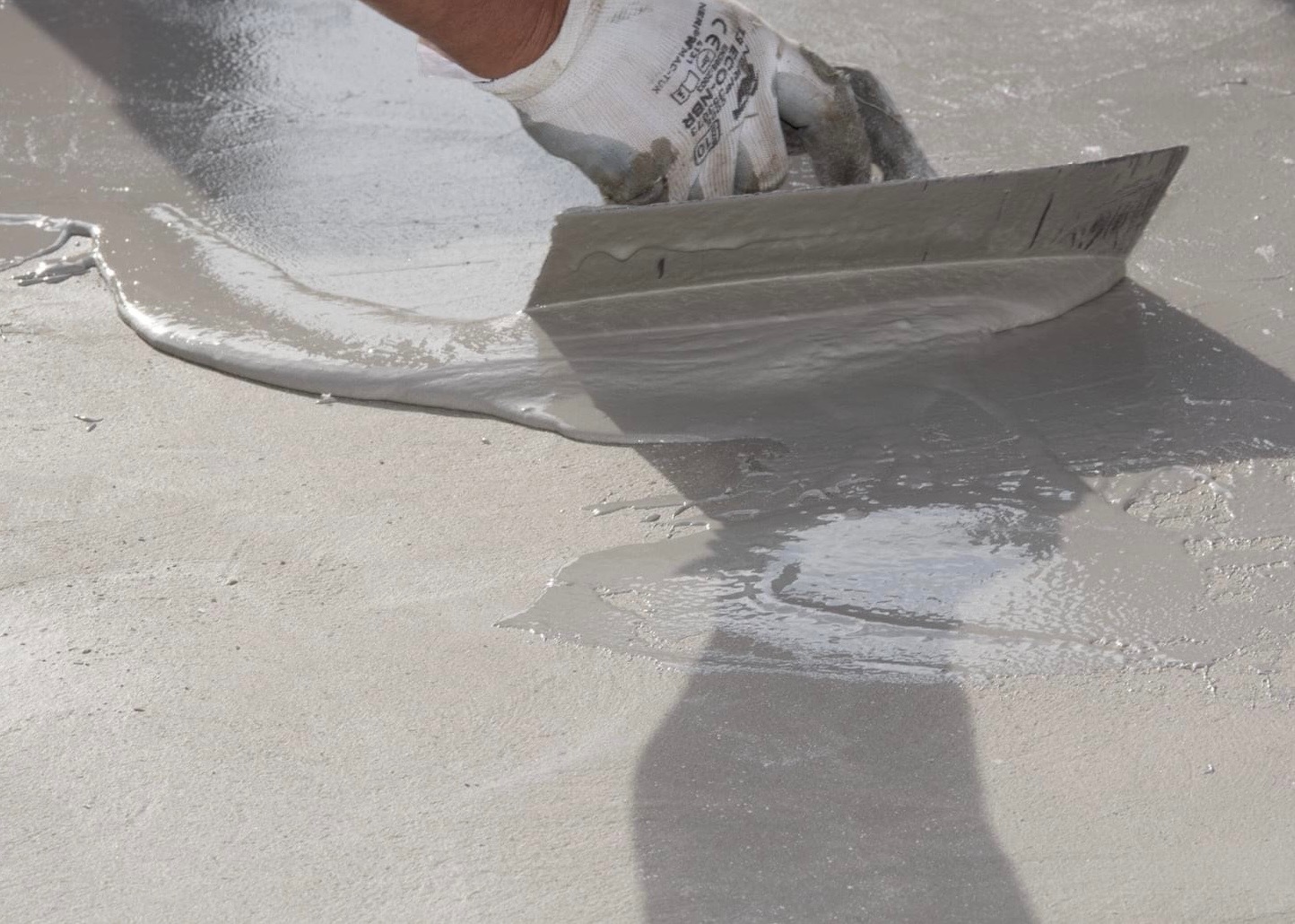 Ręka w roboczej rękawicy rozprowadza szpachlą mokrą, samopoziomującą masę na betonowej powierzchni, tworząc gładką warstwę.