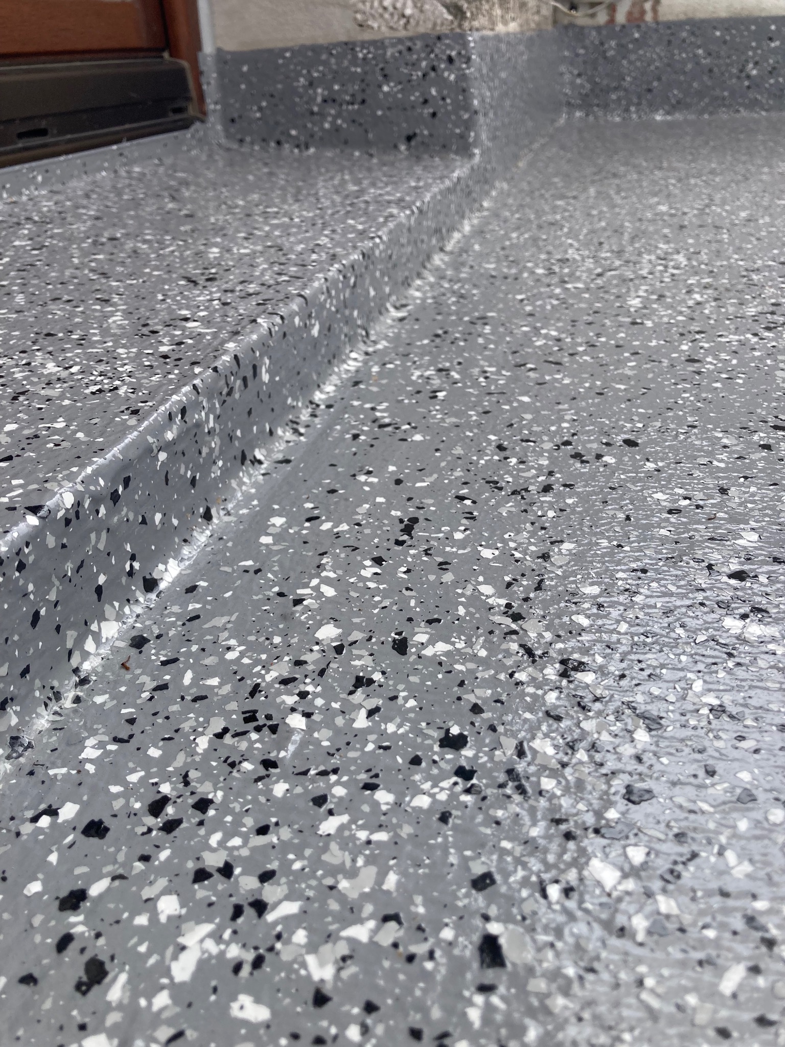 Szara, wielobarwna, żywiczna posadzka balkonu lub tarasu z widocznym progiem drzwi balkonowych i fragmentem ściany.