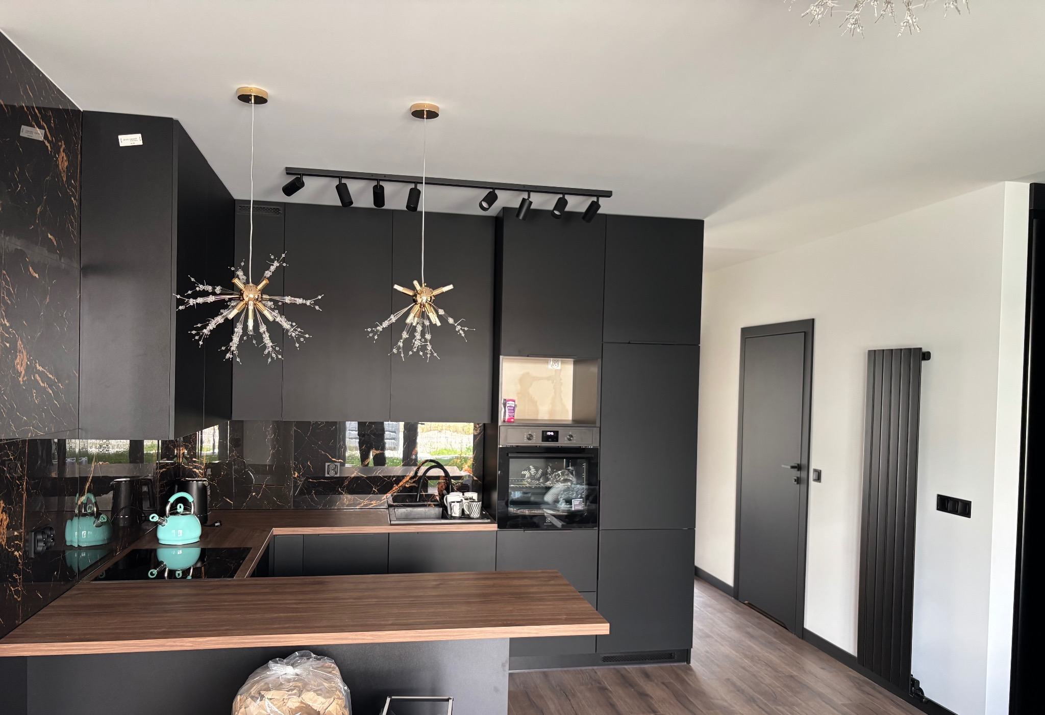 Nowoczesna kuchnia w czerni z marmurowym akcentem na ścianie i blacie. Oświetlenie punktowe i dekoracyjne lampy nad wyspą. Minimalistyczny design z elementami złota.