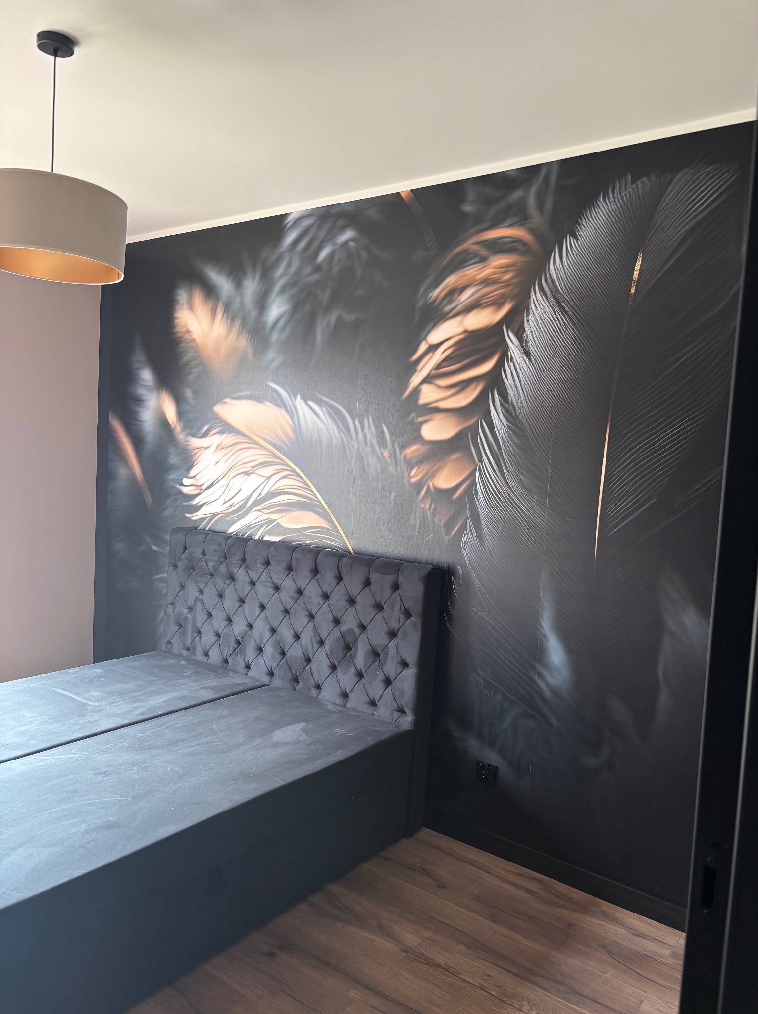 Sypialnia z łóżkiem i fototapetą z motywem piór w odcieniach szarości i złota. Na suficie widoczna lampa. Podłoga z paneli w ciepłym odcieniu brązu.