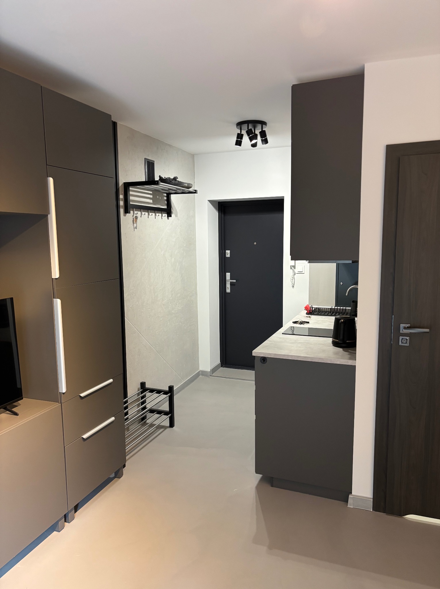 Nowoczesne wykończenie wnętrza przedpokoju z szarą zabudową meblową, wieszakiem na ubrania i butami na półce. Minimalistyczny design, neutralna kolorystyka.