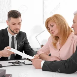 Młody mężczyzna w garniturze wręcza dokument parze siedzącej przy biurku, na którym leżą okulary, długopis i pieczątka.