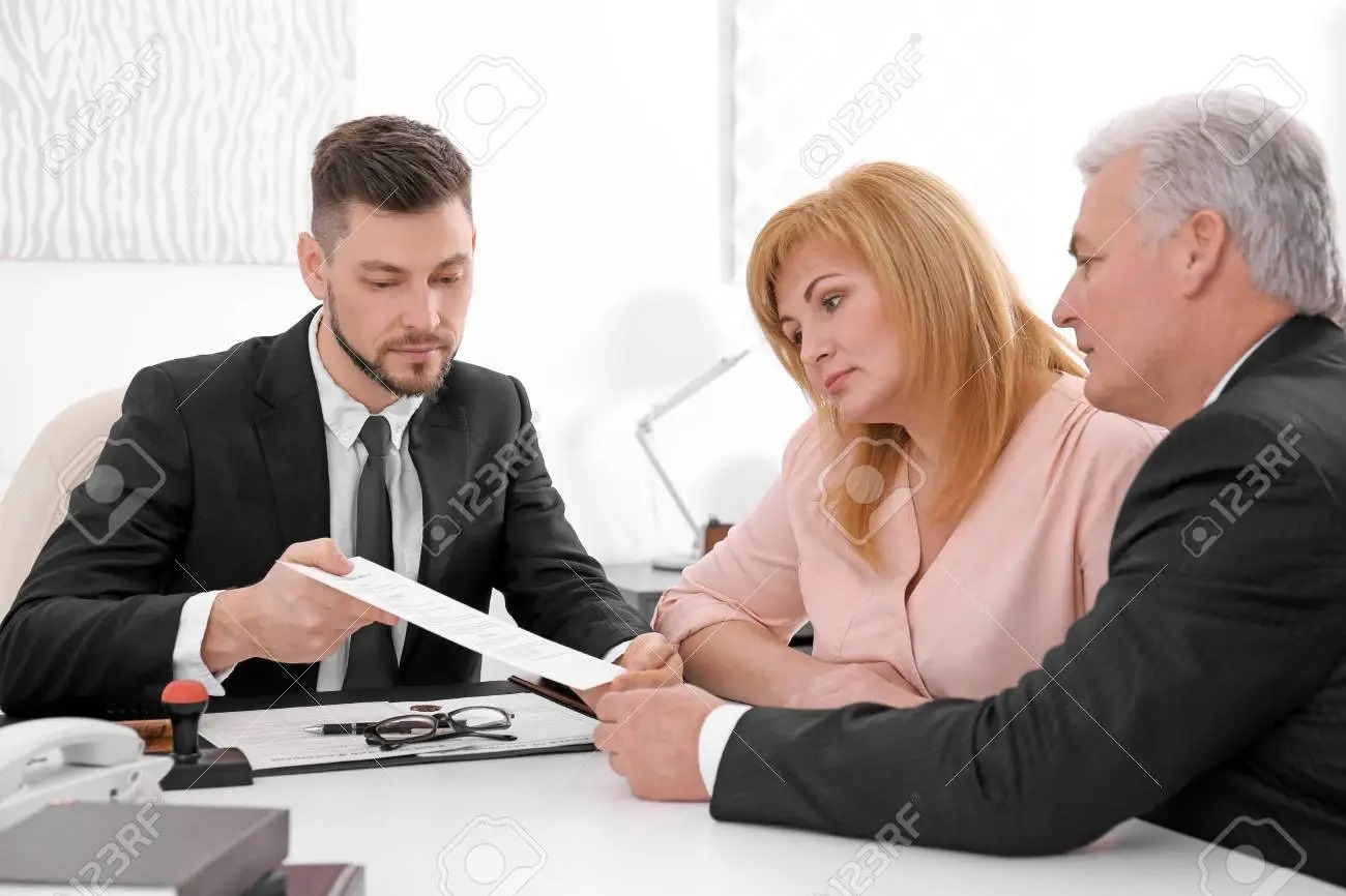 Młody mężczyzna w garniturze wręcza dokument parze siedzącej przy biurku, na którym leżą okulary, długopis i pieczątka.