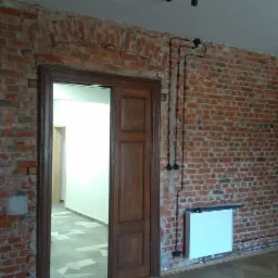 Surowa ceglana ściana z odrestaurowanym łukiem nad drewnianymi drzwiami, widoczne instalacje elektryczne natynkowe i grzejniki, podłoga parkietowa.