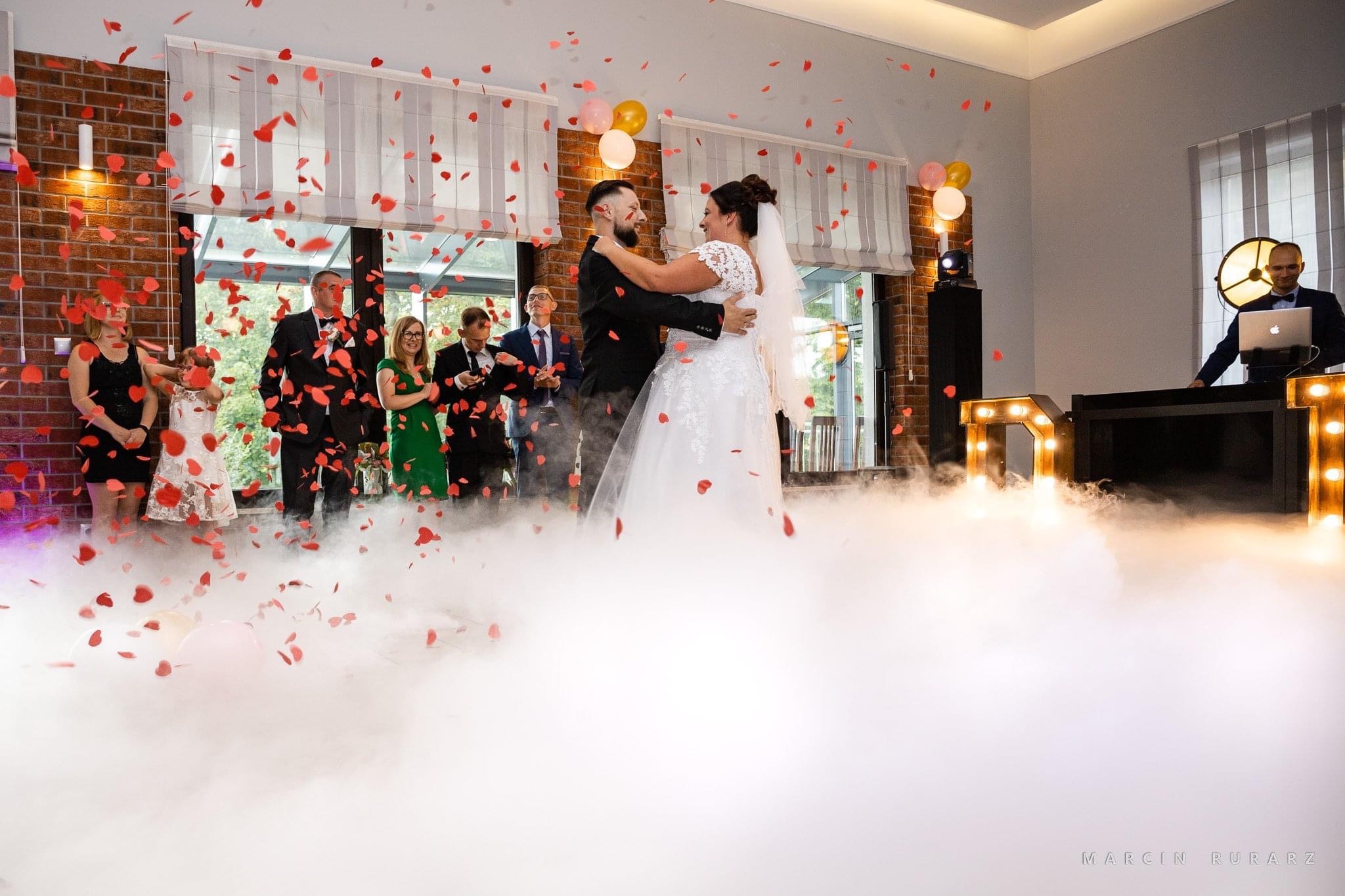 Pierwszy taniec pary młodej w otoczeniu gości, opadających płatków róż i efektu ciężkiego dymu, DJ z laptopem w tle.