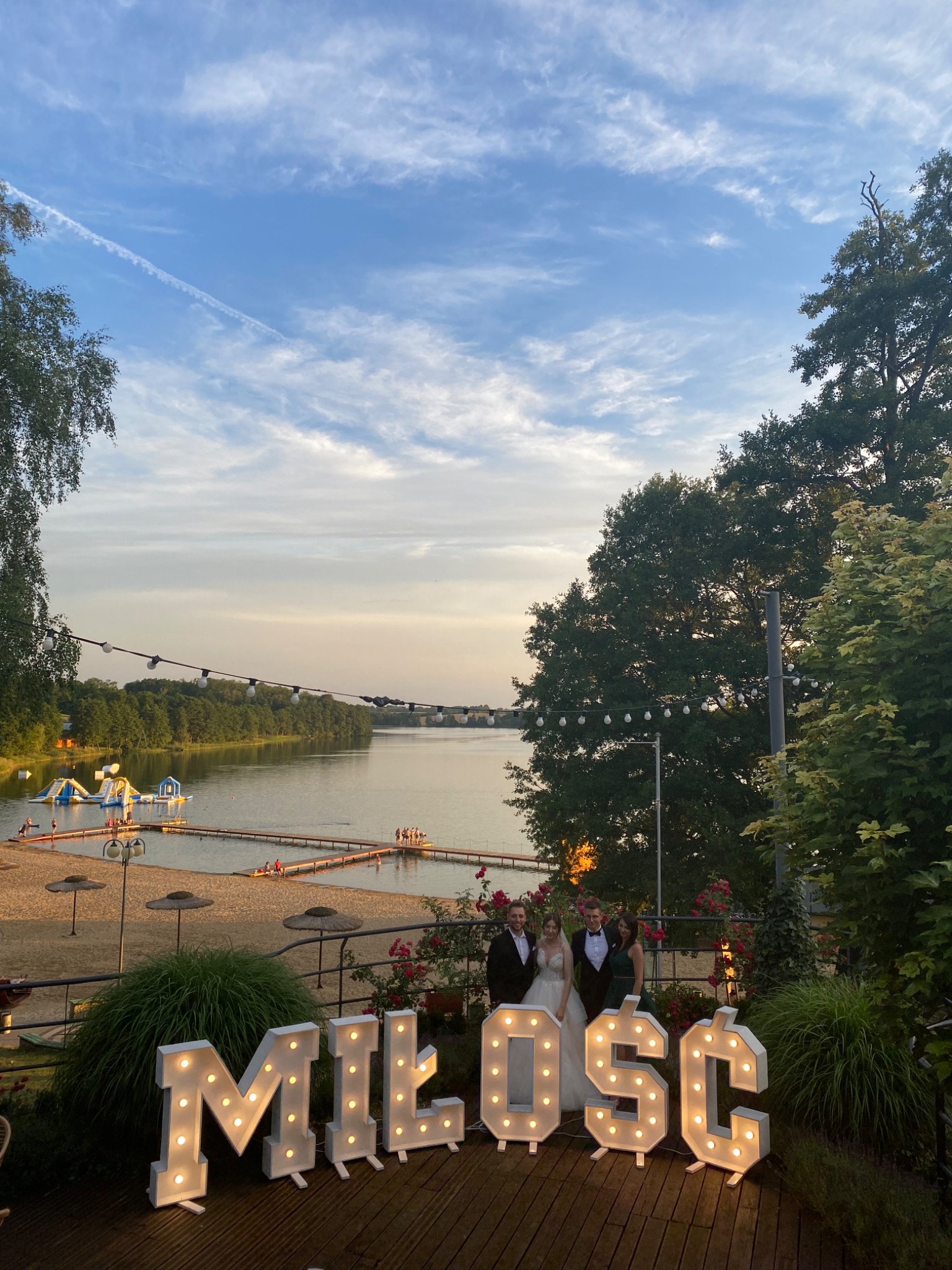 Zespół muzyczny w trakcie występu na weselu plenerowym nad jeziorem, z podświetlanym napisem 'MIŁOŚĆ' na pierwszym planie i parą młodą w centrum kadru.