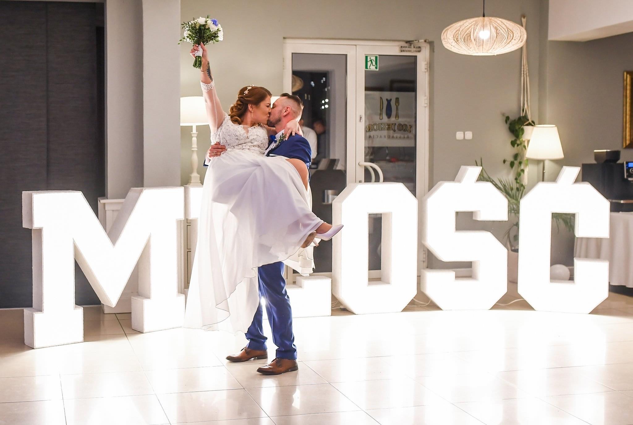 Para młoda w objęciach, panna młoda trzyma bukiet, w tle duże, podświetlane litery układające się w słowo 'MIŁOŚĆ', widoczny fragment restauracji.