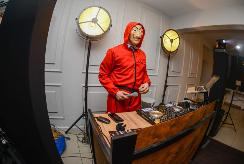 Osoba w czerwonym kombinezonie i masce Salvadora Dali stoi za konsoletą DJ-ską, trzymając mikrofon, oświetlona przez dwa reflektory sceniczne.