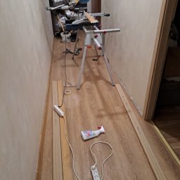 MarioBau Mariusz Górecki - Przygotowania do montażu listew przypodłogowych: ukośnica Makita na stojaku, listwy przy ścianie, przedłużacz i płyn do czyszczenia na podłodze imitującej drewno.