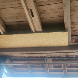 MarioBau Mariusz Górecki - Montaż płyty OSB na drewnianej konstrukcji poddasza, widoczne belki stropowe i fragment ściany, rusztowanie w dolnej części kadru.