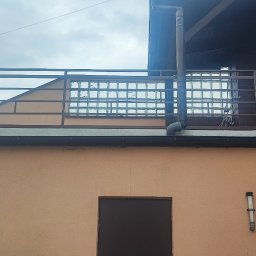 MarioBau Mariusz Górecki - Fragment budynku z balkonem z metalową balustradą, ścianą z luksferami i rynną spustową, widok z dołu na tle pochmurnego nieba.