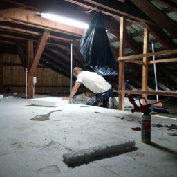 MarioBau Mariusz Górecki - Pracownik na kolanach docina białą płytę izolacyjną piłą ręczną na poddaszu z widoczną konstrukcją drewnianą, workiem foliowym oraz narzędziami pracy.