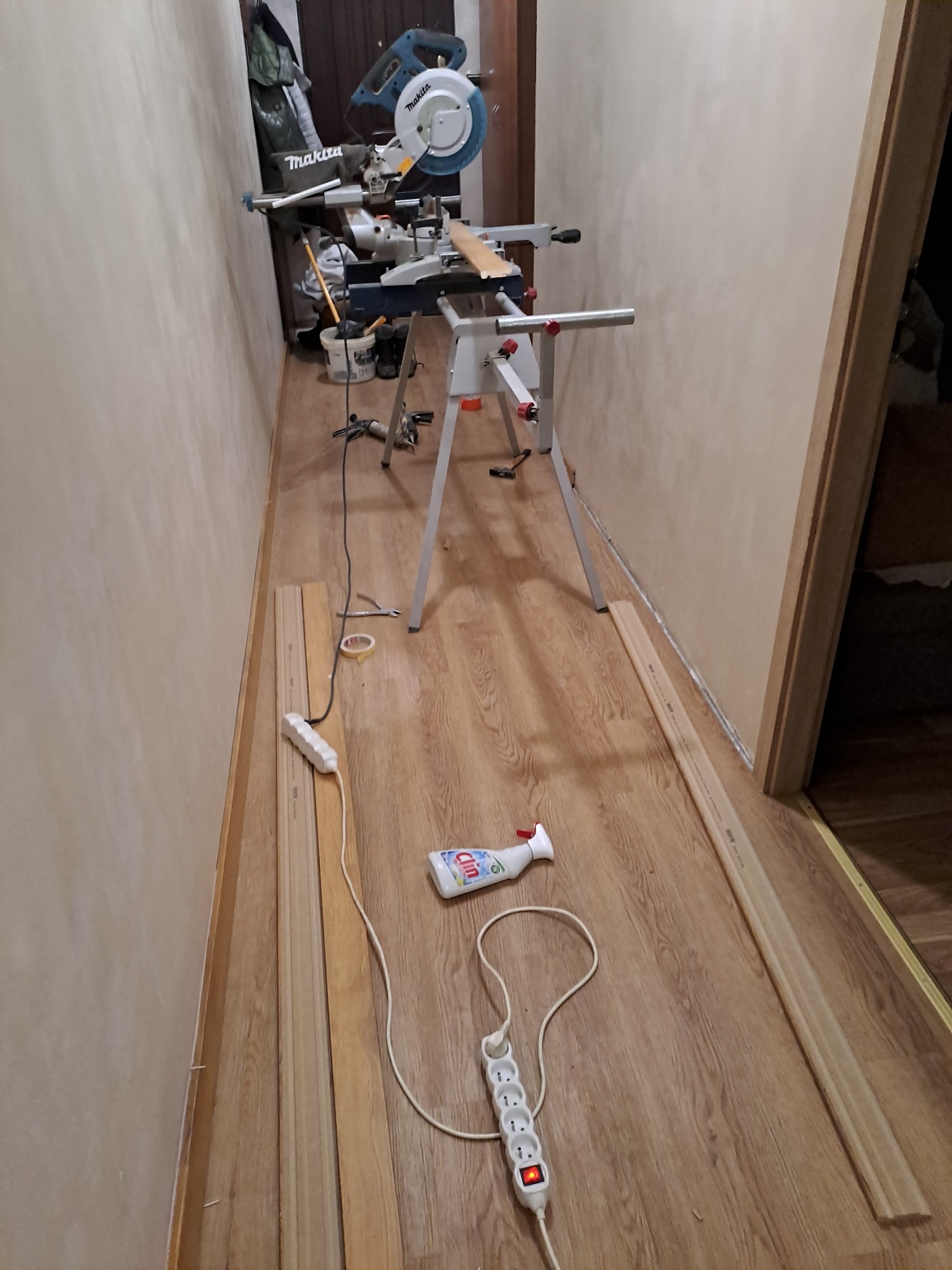 Przygotowania do montażu listew przypodłogowych: ukośnica Makita na stojaku, listwy przy ścianie, przedłużacz i płyn do czyszczenia na podłodze imitującej drewno.