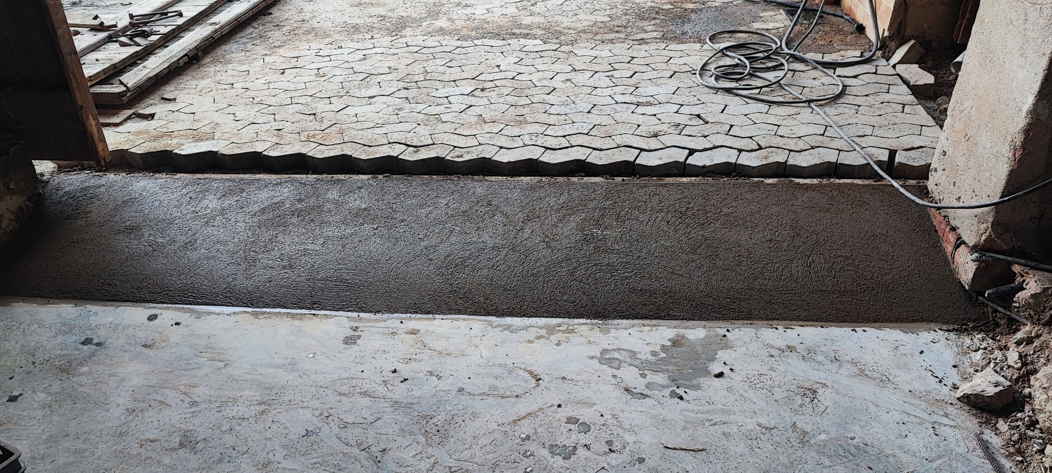 Fragment podjazdu z ułożoną częścią kostki brukowej w kształcie rombów i świeżo wylaną, mokrą zaprawą cementową, widoczne narzędzia i kable elektryczne.