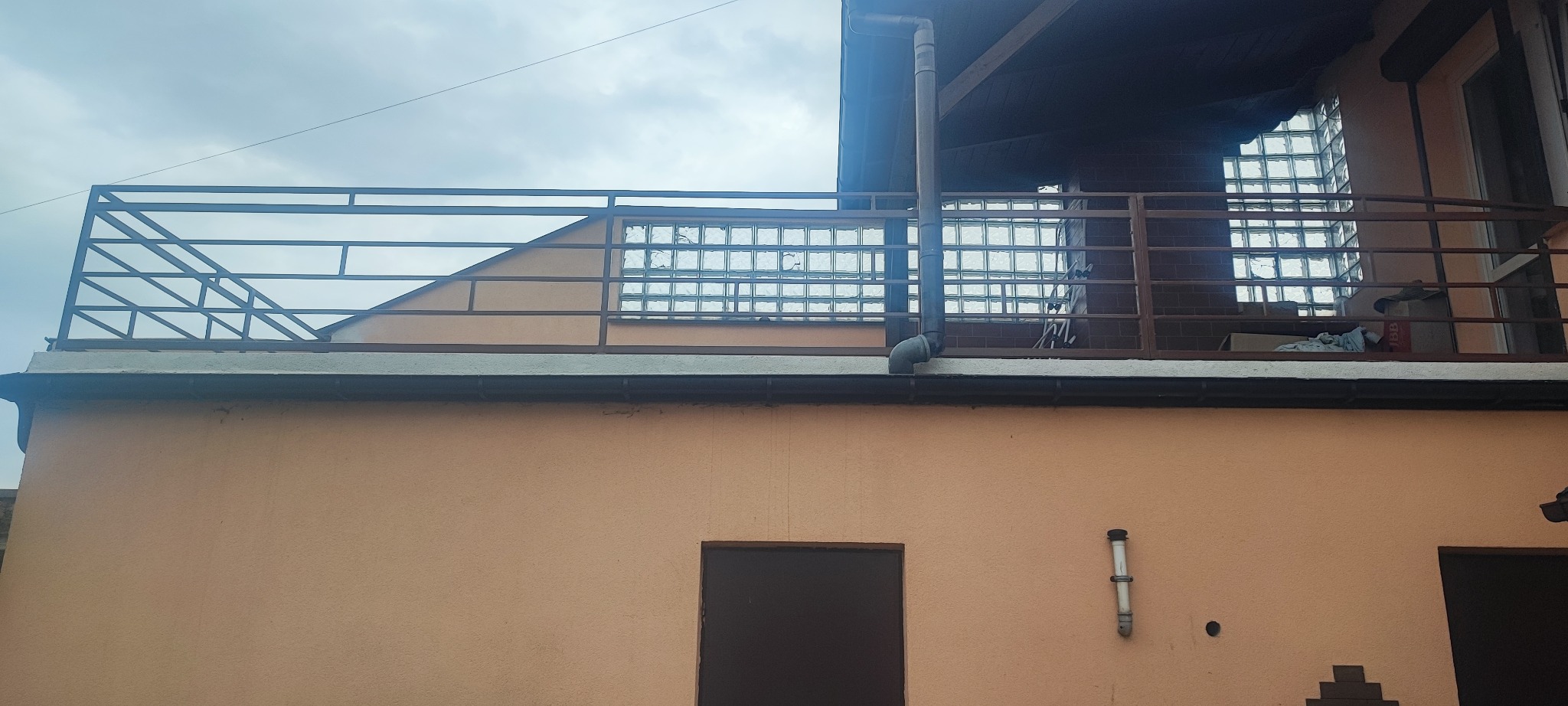 Fragment budynku z balkonem z metalową balustradą, ścianą z luksferami i rynną spustową, widok z dołu na tle pochmurnego nieba.
