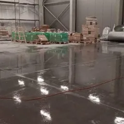 Błyszcząca, mokra posadzka przemysłowa w hali magazynowej po umyciu, z widocznymi paletami i materiałami budowlanymi w tle.