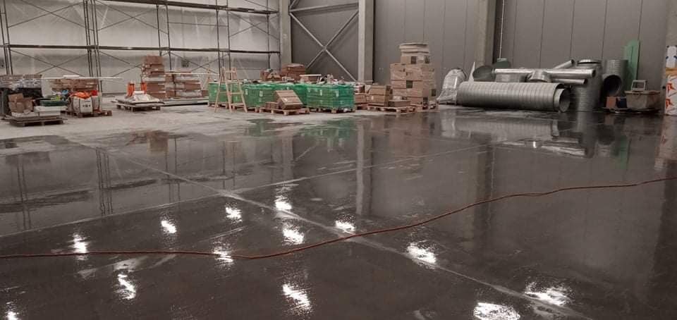 Błyszcząca, mokra posadzka przemysłowa w hali magazynowej po umyciu, z widocznymi paletami i materiałami budowlanymi w tle.