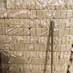 Etna Zbigniew Przepiórski - Stos ułożonych, zapakowanych w folię paczek brykietu drzewnego z widocznym drewnianym klockiem z boku i kijem opartym o stos.