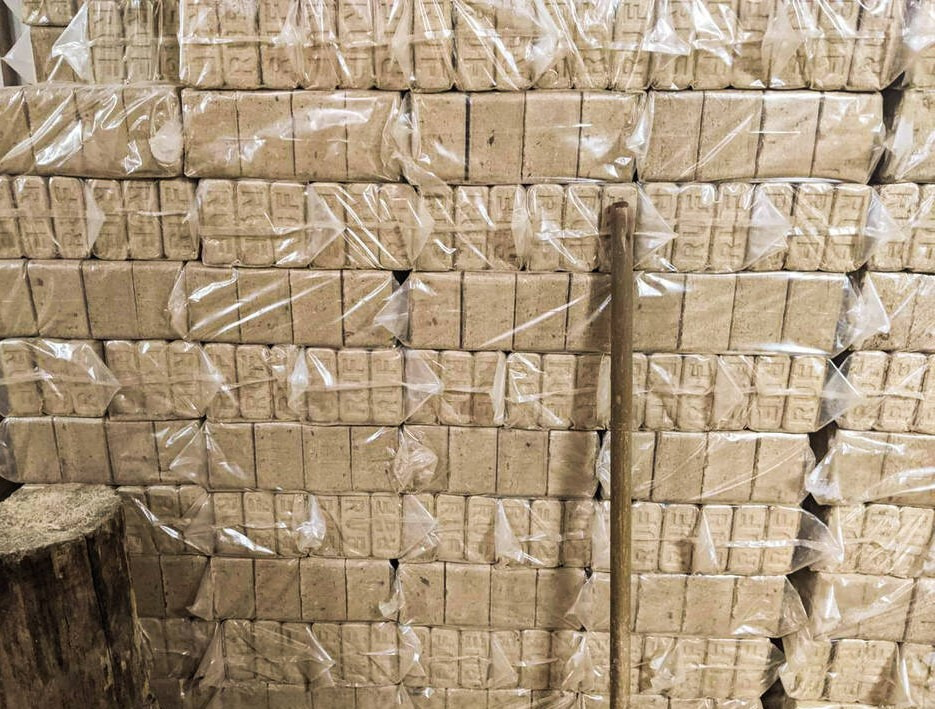 Stos ułożonych, zapakowanych w folię paczek brykietu drzewnego z widocznym drewnianym klockiem z boku i kijem opartym o stos.