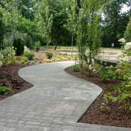 Kręty chodnik z szarej kostki brukowej prowadzący przez ogród z rabatami, wyściółkowanymi korą, z różnorodną roślinnością, w tle drzewa.