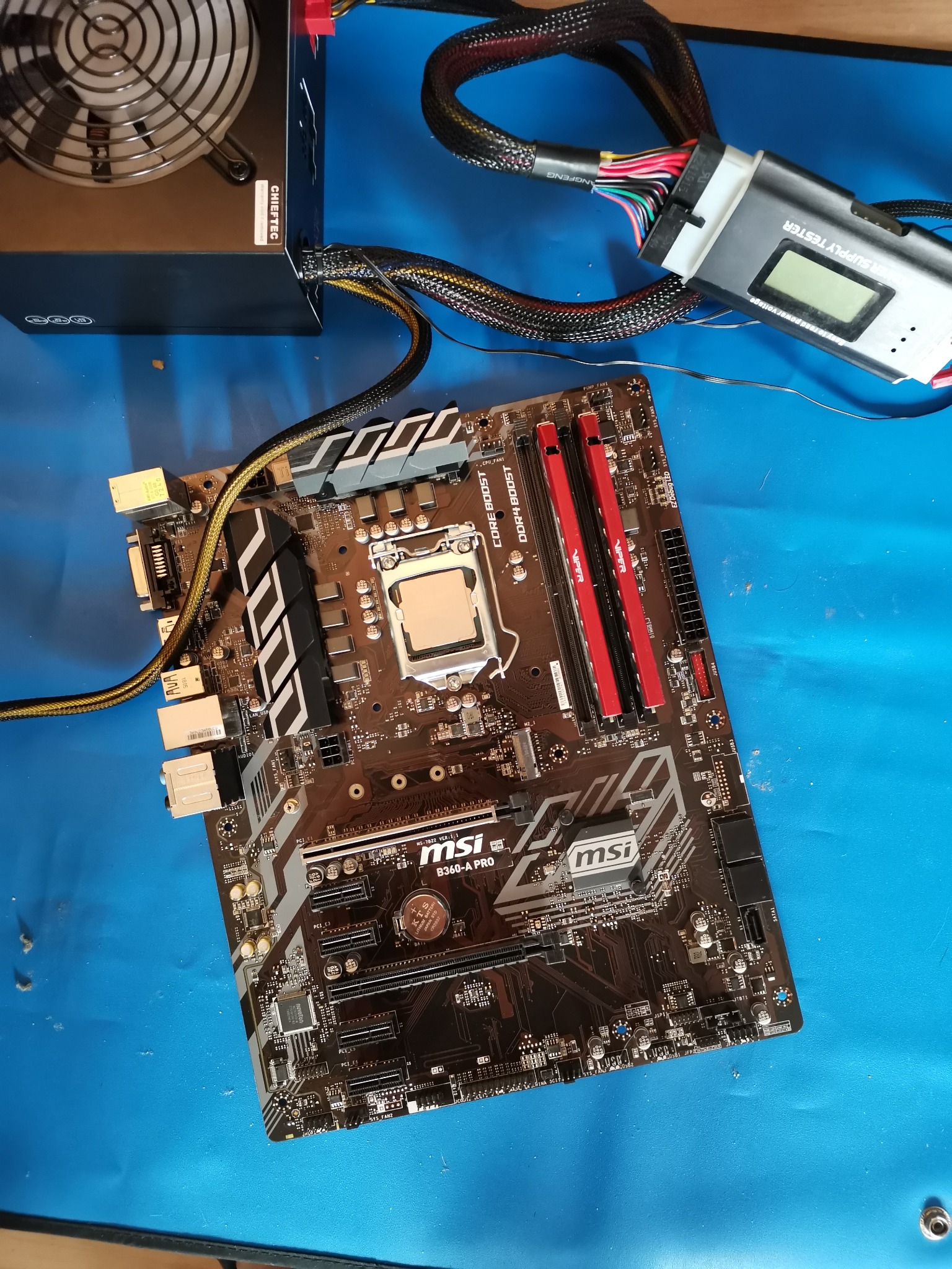Płyta główna MSI B360-A PRO z zainstalowanym procesorem i pamięcią RAM, zasilacz Chiefttec i tester zasilaczy na niebieskim podłożu.
