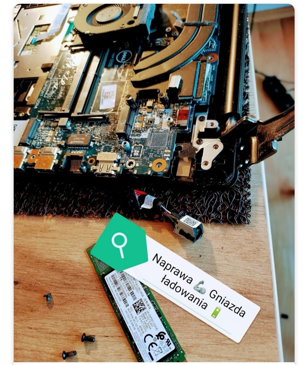 Wnętrze otwartego laptopa Dell z widoczną płytą główną, wentylatorem i złączami, obok leży dysk SSD M.2 Samsung z naklejką informacyjną, śrubki oraz kartka z napisem 'Naprawa gniazda ładowania'.