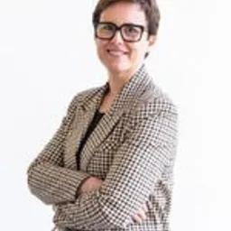 Uśmiechnięta kobieta w okularach, ubrana w brązowo-białą marynarkę w pepitkę, pozuje na białym tle z założonymi rękami.