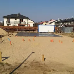 Przygotowany teren pod budowę domu jednorodzinnego z wylaną płytą fundamentową, widoczne wystające rury kanalizacyjne, zwój niebieskich rur oraz kontener budowlany w tle.