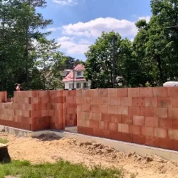 Wznoszone mury z czerwonej cegły na fundamencie, widoczne drzewa i dom w tle, piasek na ziemi, fragment ławki z pniaków.