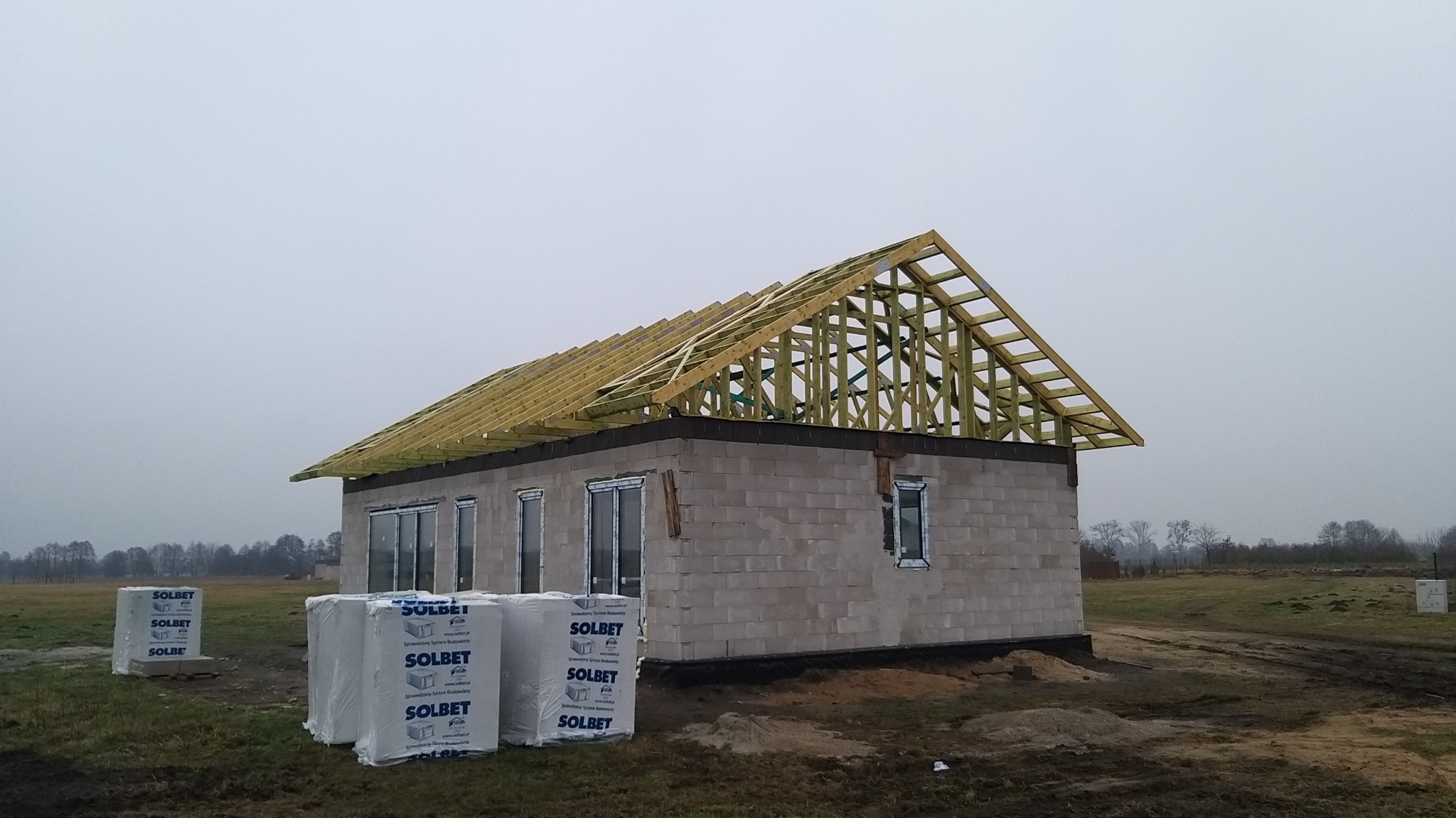 Surowy stan domu z szarej cegły z drewnianą konstrukcją dachu, widoczne okna i palety cegieł Solbet na tle pochmurnego nieba i pola.