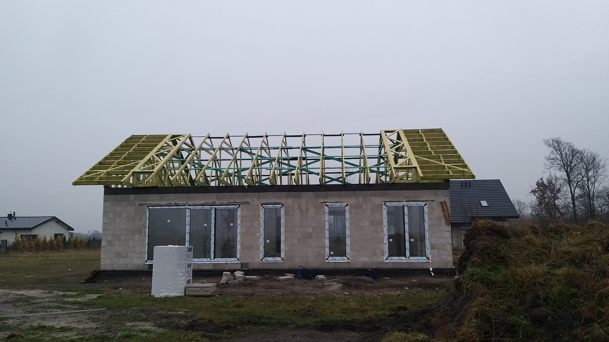 Niedokończony parterowy dom z szarego bloczka z zamontowanymi oknami i drewnianą konstrukcją dachu, widok z przodu w pochmurny dzień.